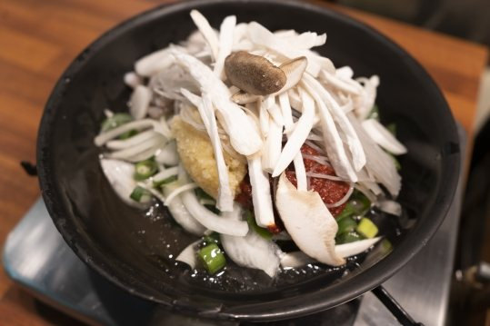 가스버너 위 검은 냄비에 담긴 익히기 전 재료 — 흰 채 썬 재료·버섯·양파·대파와 붉은 양념